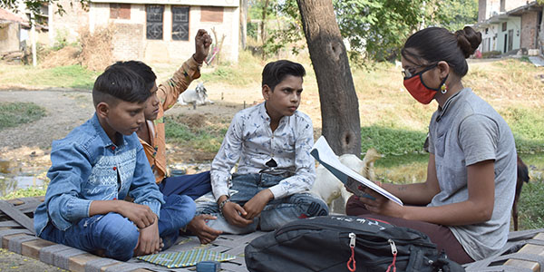 A teenage girl, wearing eyeglasses, tutors three boys in her village.