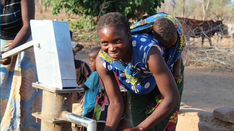 A mother, carrying her baby on her back, happily collects water from the borehole