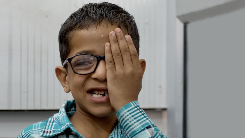 A young boy, wearing eyeglasses, covers his left eye with his left hand and smiles at the camera