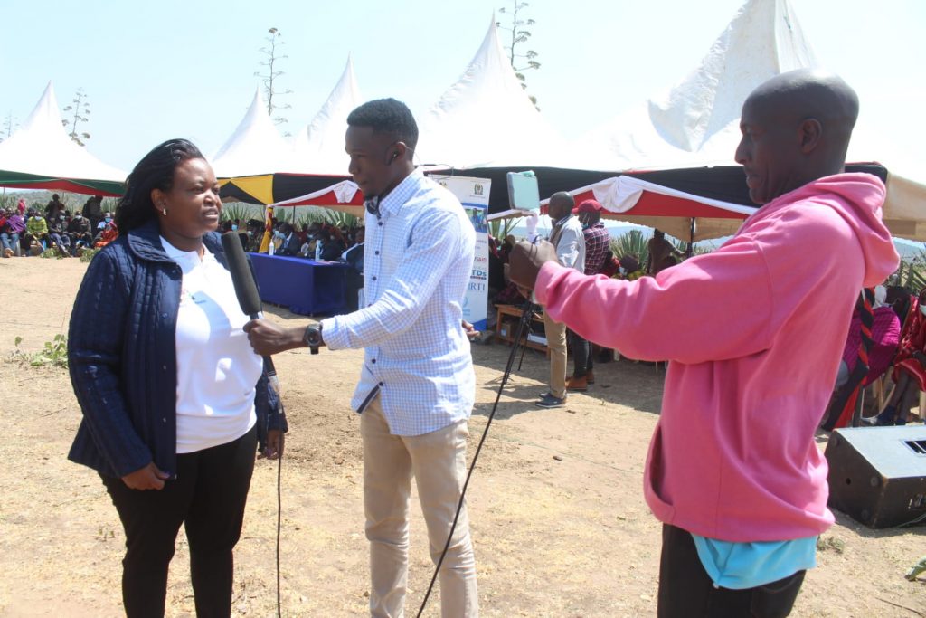 Alice, the Country Director of Operation Eyesight in Kenya, is interviewed. Her interviewers hold a mobile phone camera and mic as they record the interview. In the background is a community gathered together.