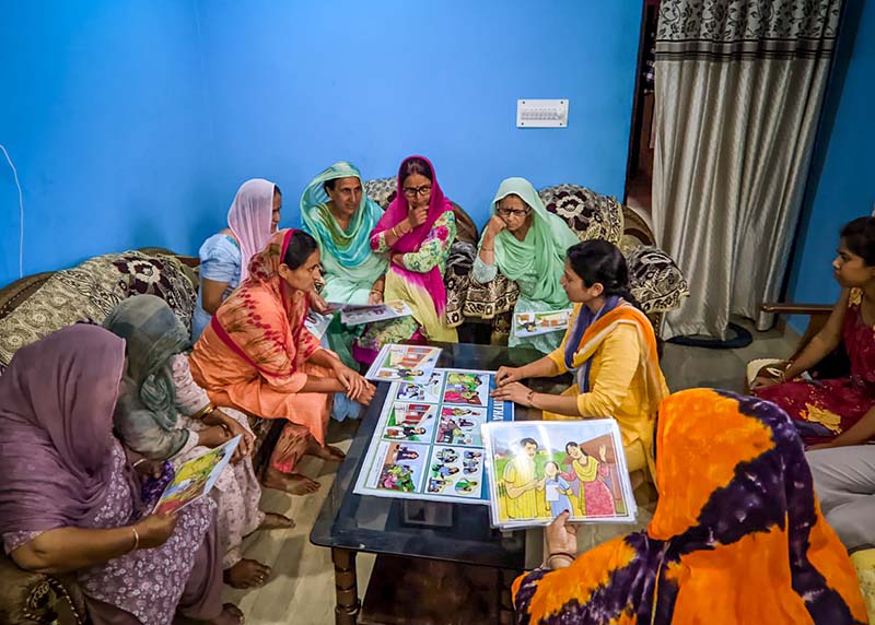 A group of women gather around educational materials in a room with blue walls.
