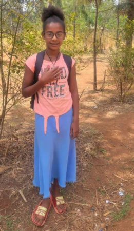 A teenaged girl stands posed for a photo in a wooded area, wearing a long skirt, t-shirt and eyeglasses.