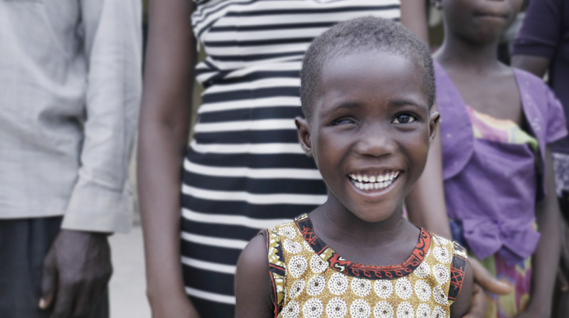A photo of a child with visual impairment smiles at the camera with other members of her community standing behind her.