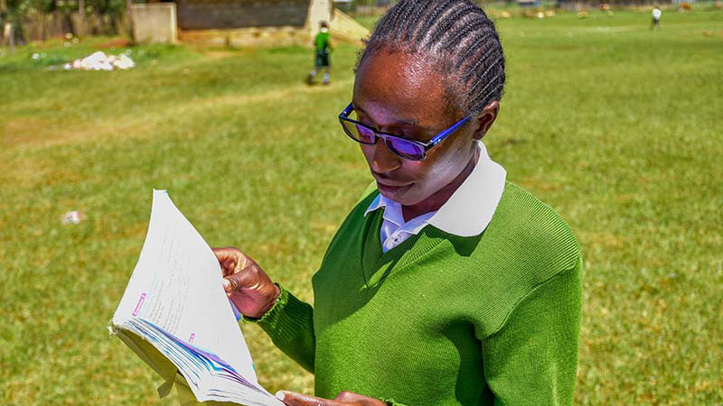A teenaged girl in a green sweater and blue eyeglasses stands reading a book in a schoolyard.