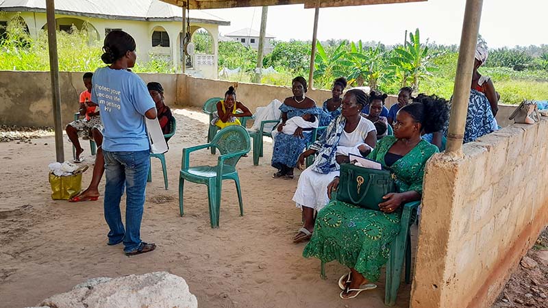 A woman holds up an eye chart to people to people not seen in the photo. Behind her, you can see sandy terrain, palm trees and a fishing boat with several men on board.