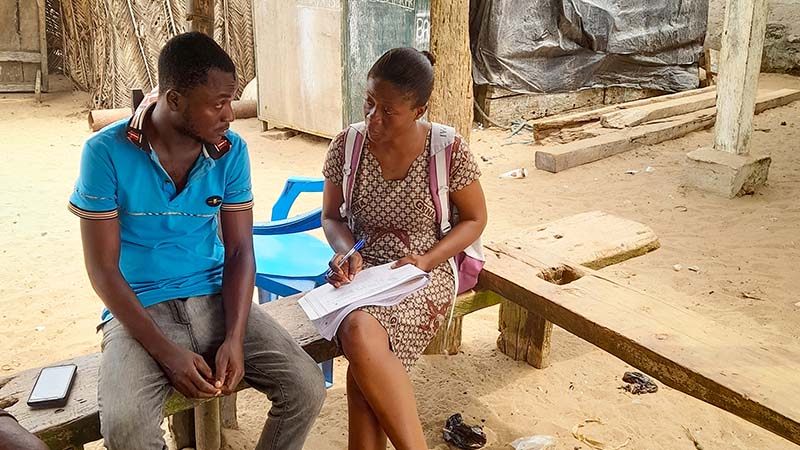 A woman interviews a man, taking notes on a clipboard. They sit outdoors on a bench in a rural village setting.