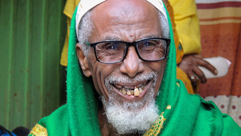 An old man with white beard, wearing skull cap and black glasses, grins at the camera.