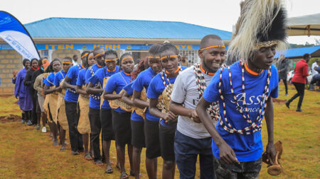 People line up in formation, dancing in front of a building with a blue roof.