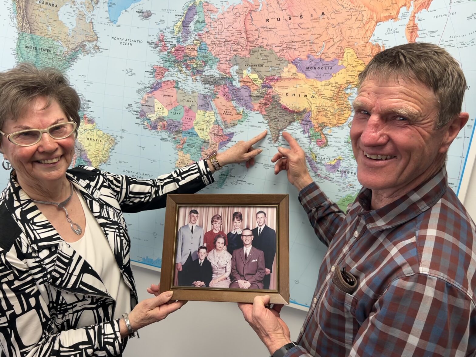 Man and woman stand in front of a world map, both pointing to the country India on the map. Together they are holding a family picture.