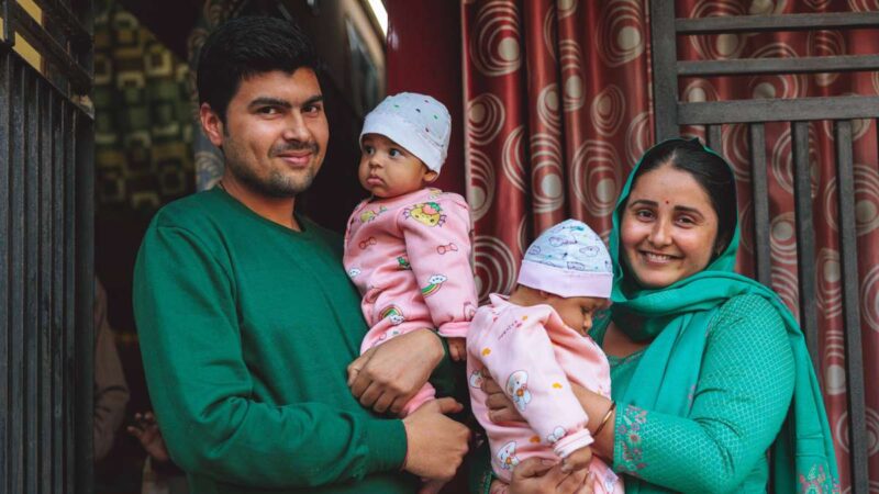 A man and his wife stand holding twin baby girls dressed identically in pink onesies.