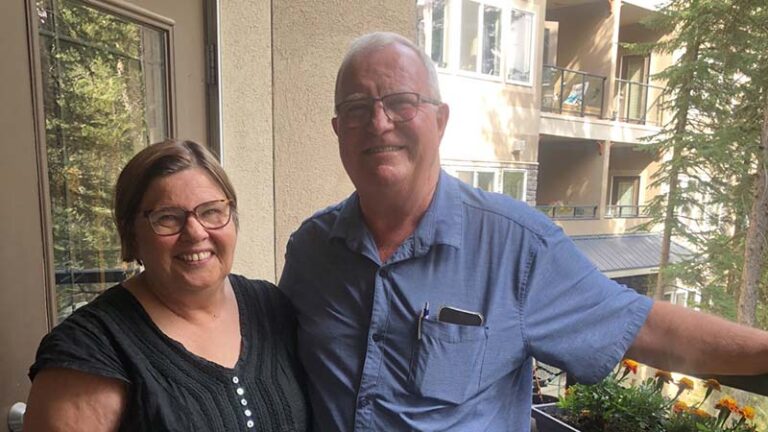 A husband and wife stand, smiling, with arms around each other.