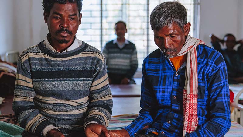 Man sits on a hospital bed, with with elderly father sitting beside him, holding his son's hand.