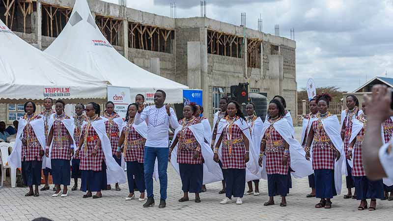 A line of people wearing white shawls and red checkered dresses dance in front a semi-constructed building. 