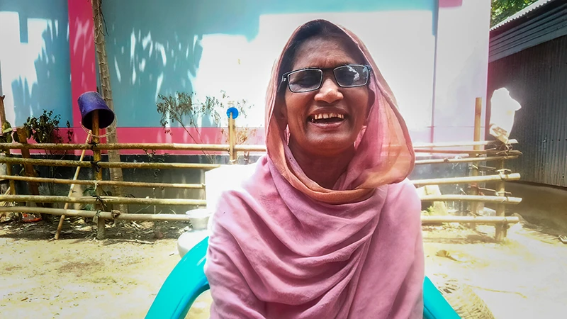 A woman wearing glasses smiles for the camera while seated in front of her home.