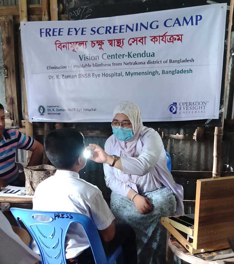 Woman shines a light into the eyes of a male student as part of an eye health screening. Behind them is a banner advertising an eye screening camp. 