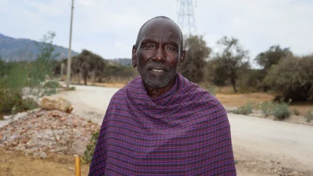 A man wearing a plaid wrap poses for a photo outside. You can see a dirt road, trees and part of a hillside in the background.