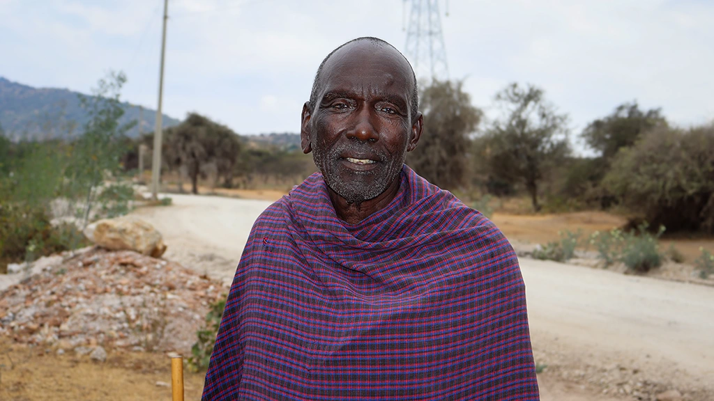 A man wearing a plaid wrap poses for a photo outside. You can see a dirt road, trees and part of a hillside in the background.
