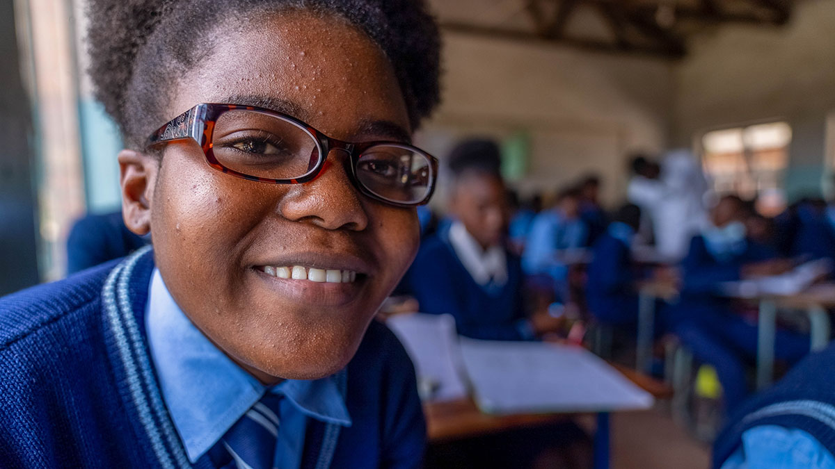 A teenaged girl wearing eyeglasses smiles at camera in classroom.