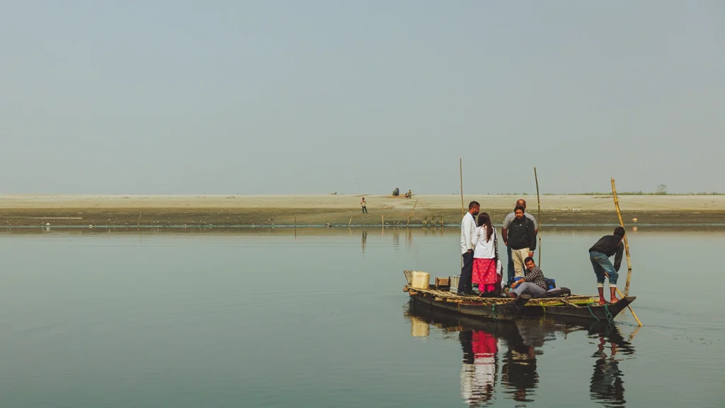 A small boat carrying a half dozen people crosses a river. The people are travelling to remote villages on a river island in India.