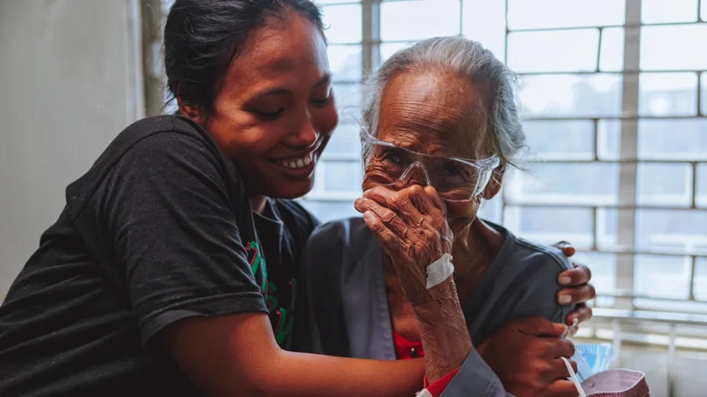 A young woman hugs a white-haired woman. The older woman wears a hospital gown and protective, and appears to be wiping away tears.