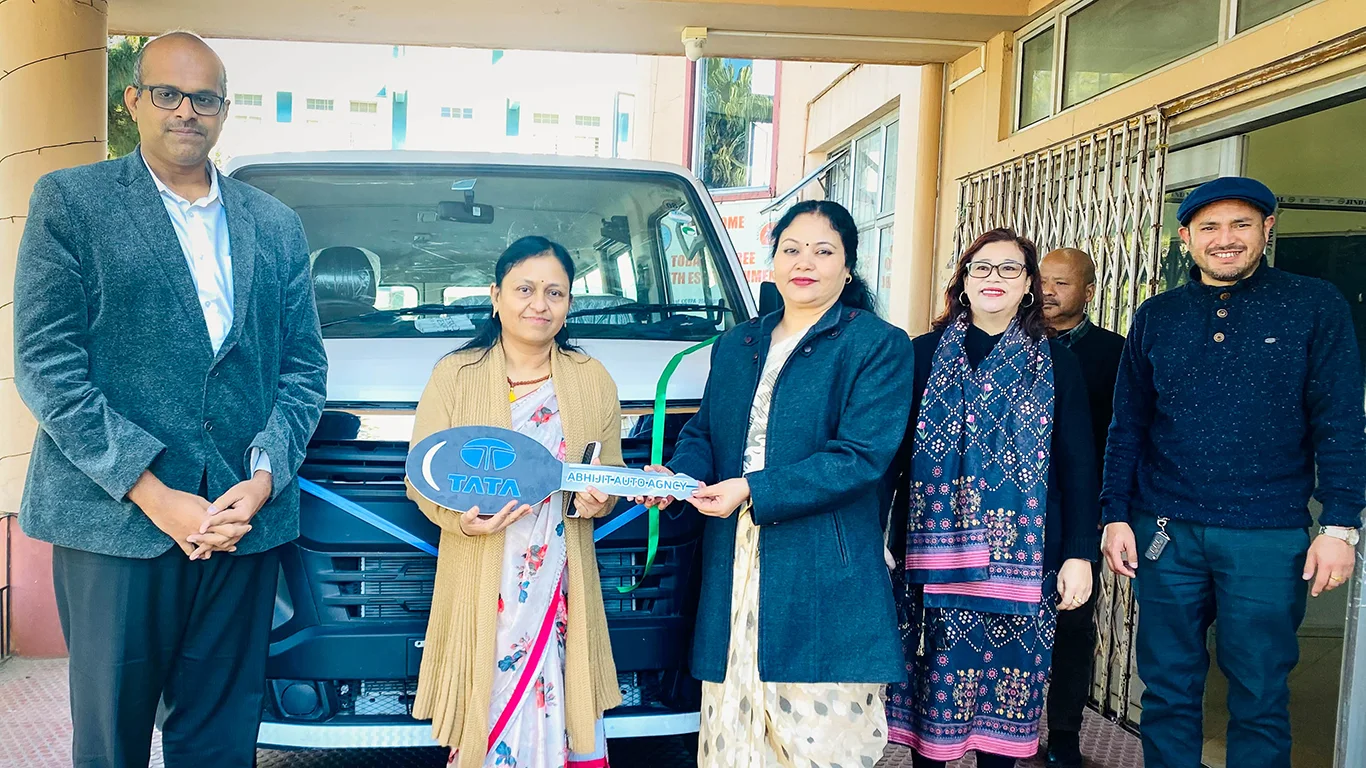 A woman hands over a large key to another woman. They stand in front of a van, surrounded by four other men and women.