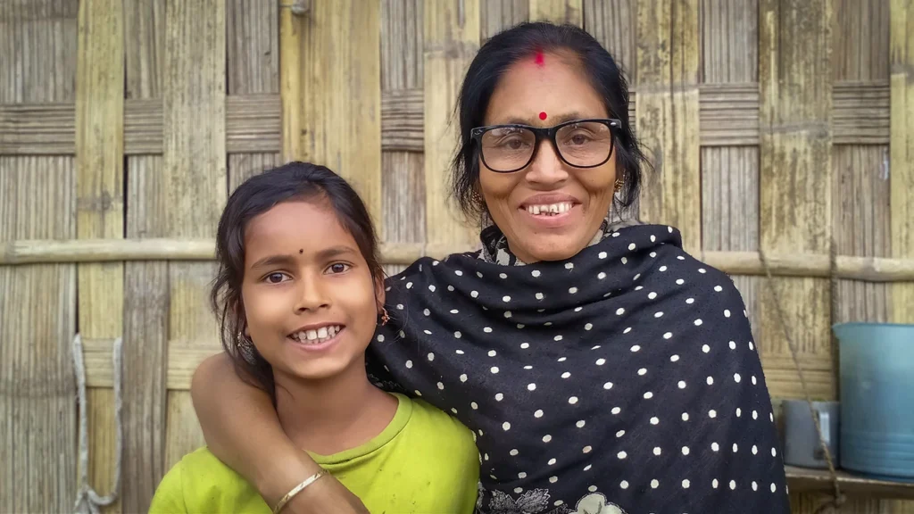 A woman wearing black eyeglasses sits next to a young girl, her arm around the girl. They are smiling.