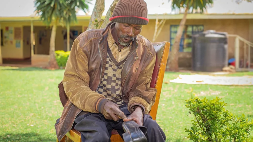 A man sits outside on a chair, repairing a men's leather shoe. 
