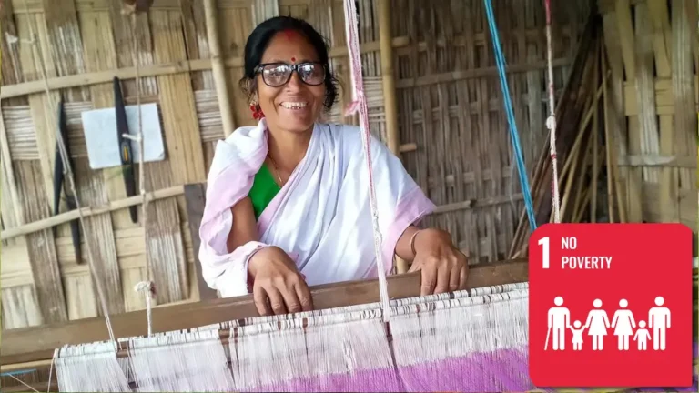 A smiling woman sits behind a hand loom. She wears eyeglasses.