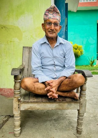 A man wearing a pair of wire-rimmed eyeglasses sits cross-legged in a wooden chair.