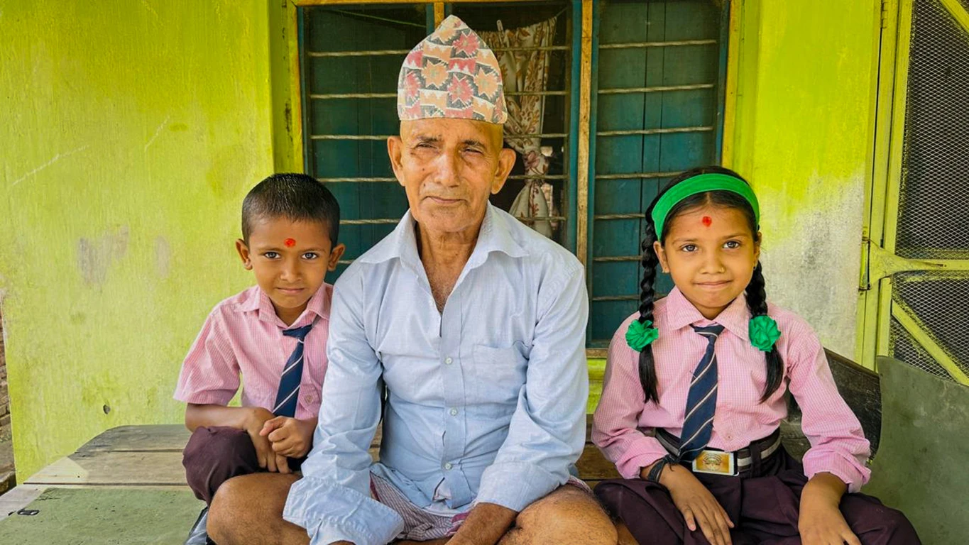 A man wearing a Nepali topi hat poses with two young children in school uniforms.