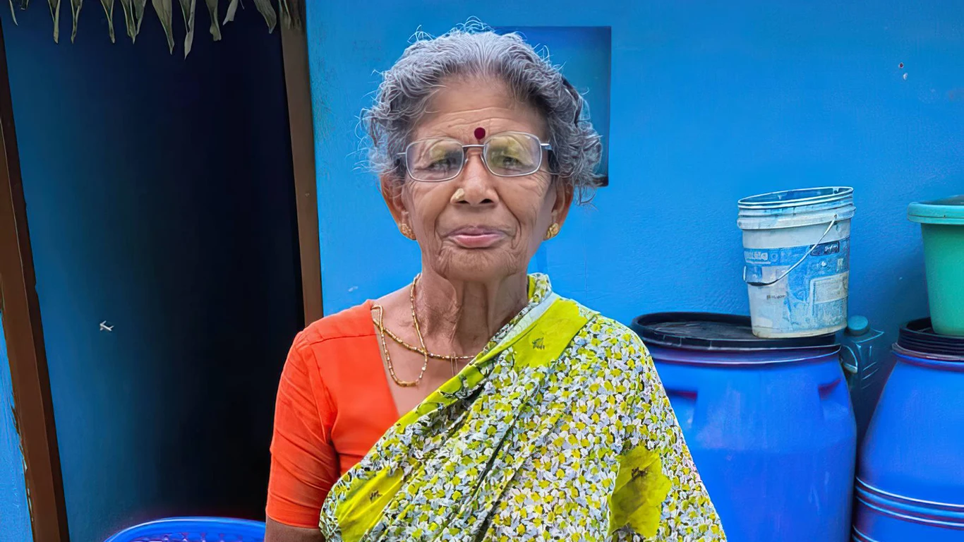 A woman stands in front of a blue house. She's wearing glasses.