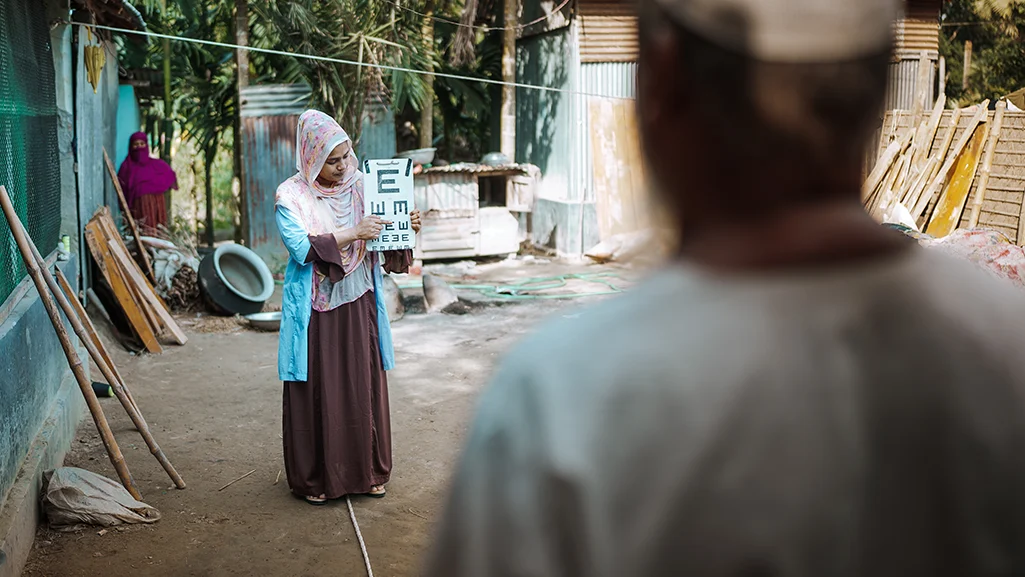 A woman holds up an eye chart to a man. They are standing outside of a family home.