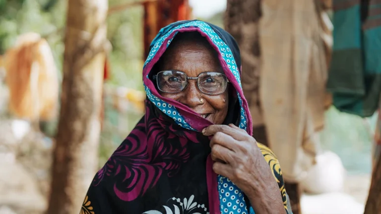 A woman wearing eyeglasses poses for a picture, partly concealing her smile with her headscarf.