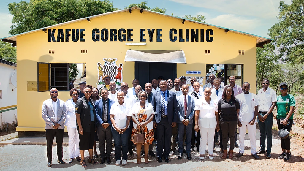 A group of people stand in front of a small yellow building with the words "Kafue Gorge Eye Clinic" painted across the front.