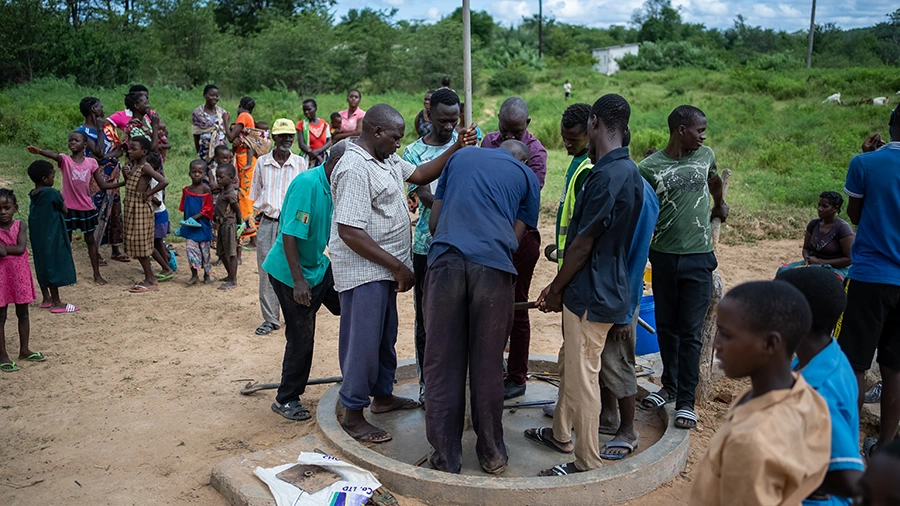 A group of men fix a borehole handpump while community members watch.