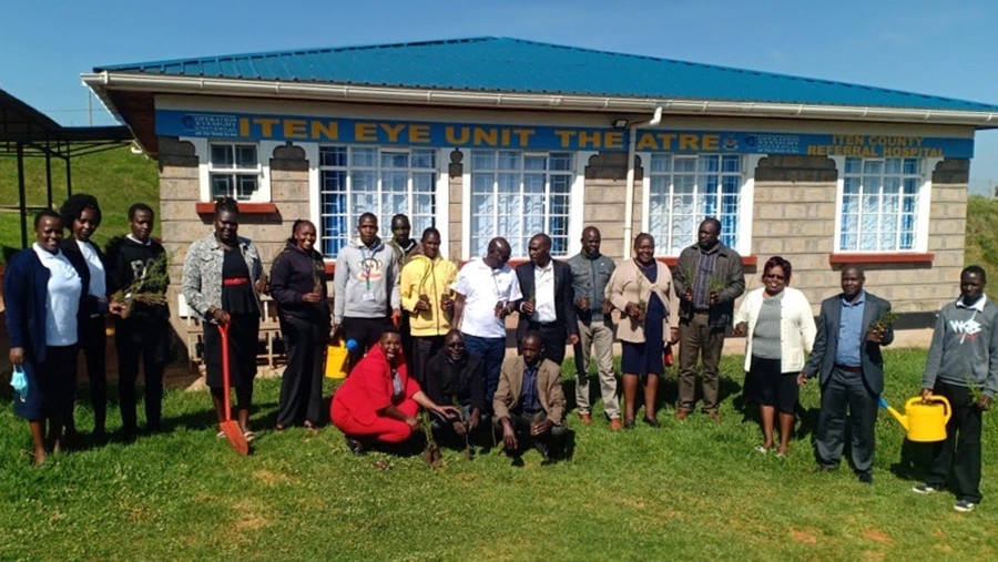 Men and women stand outside of a small eye clinic, holding shovels, seedlings and a watering can.  