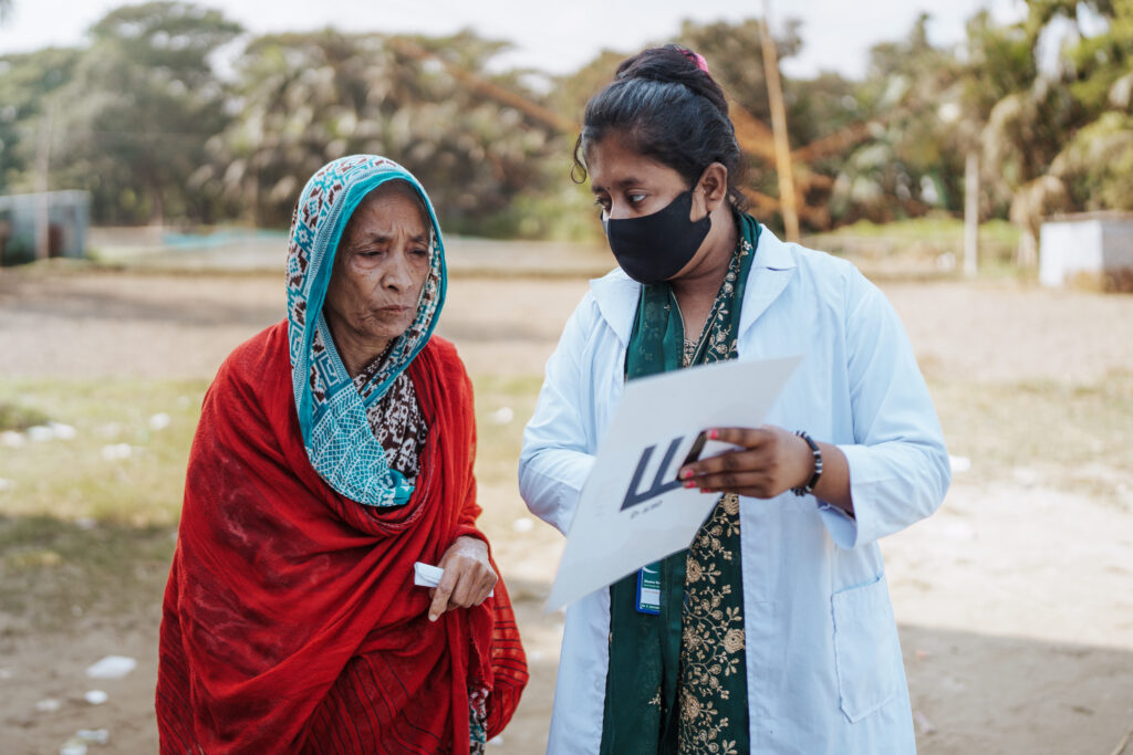 A community health worker called Jhuma Rani in a white coat asks Gulabahr about her eyes. The exam was part of a screening that led to the cataract surgery referral at Symbiosis Hospital, one of the partner hospitals of Operation Eyesight.