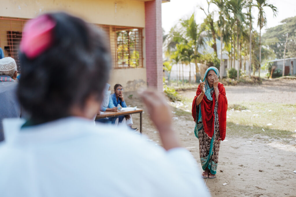 Gulbahar stands at a distance with one hand covering her left eye, during a vision screening. A community health worker in a white coat is conducting the exam at the Operation Eyesight outreach eye camp. Other individuals sit nearby outside a yellow building, with palm trees and greenery in the background.