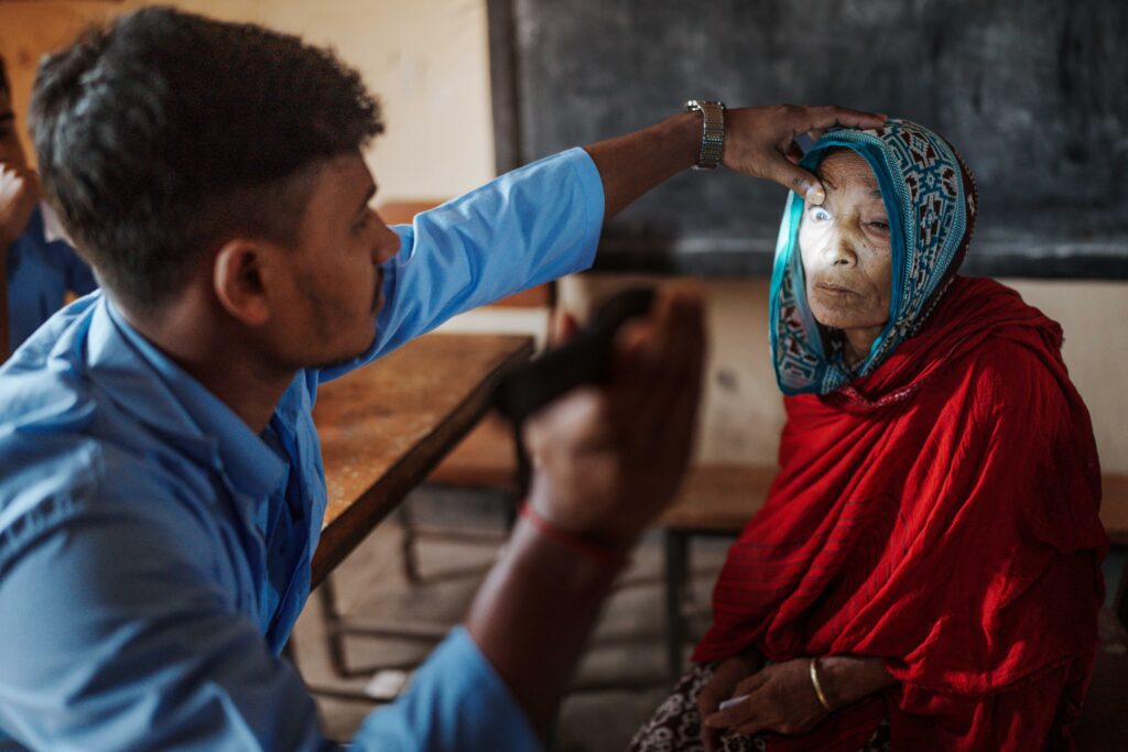 A health worker in a blue shirt conducts an eye examination on Gulbahar. They are seated in a room at the Operation Eyesight outreach eye camp with wooden desks and chairs in the background. The exam was part of a screening that led to the cataract surgery referral at Symbiosis Hospital, one of the partner hospitals of Operation Eyesight.