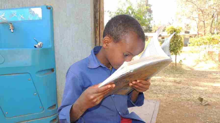 Fassikaw in a blue shirt sits on a bench outdoors, holding a book very close to his face while reading, indicating difficulty seeing clearly.
