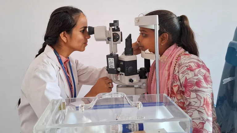 A woman in a lab coat examines the eyes of another woman through a slit lamp.
