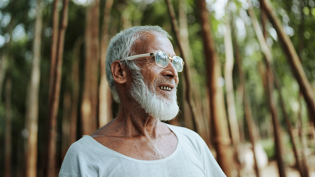 A man with white hair and beard, and wearing glasses, stands outside in a forested area, smiling.