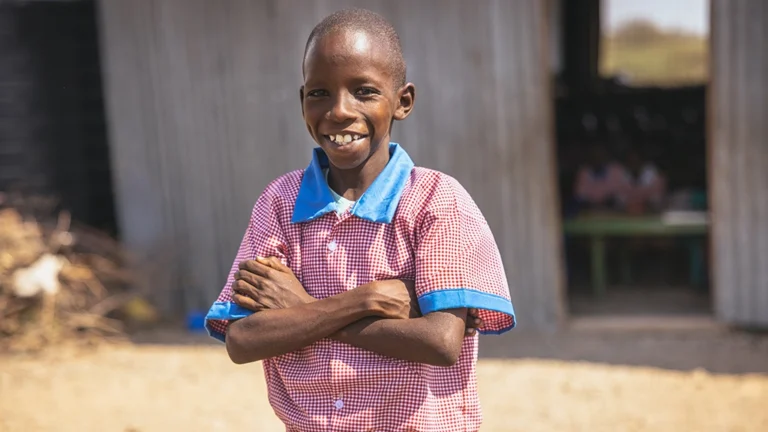 A boy in a blue and red school uniform shirt stands with his arms crossed, smiling.