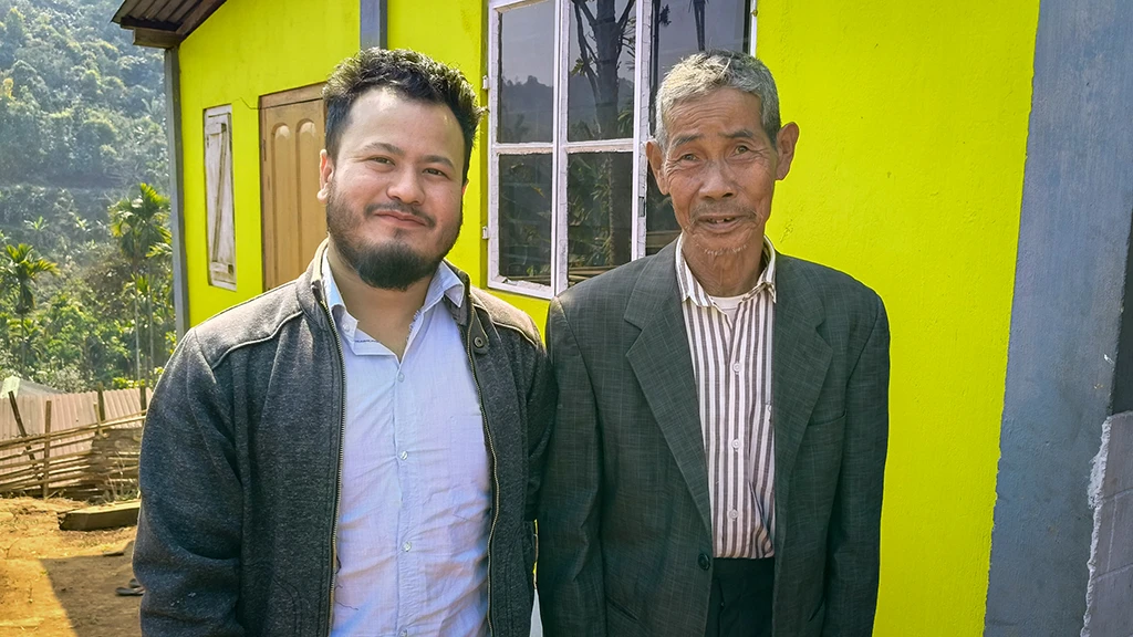 A young man stands next to an older man outside of a yellow house.